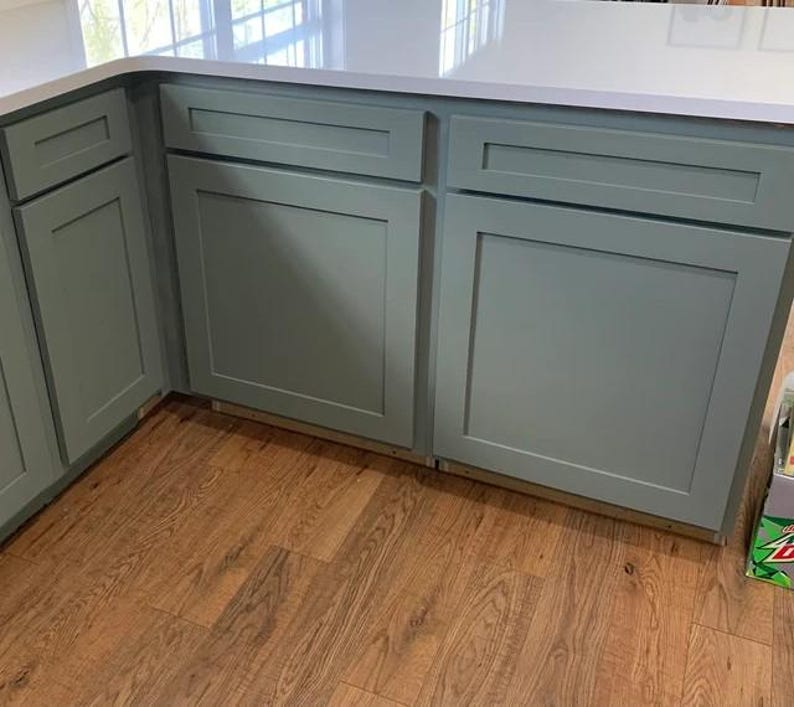May include: Sage green kitchen cabinetry featuring a shaker-style design with flat recessed center panels and horizontal drawer fronts. The cabinets are topped with a smooth white countertop and installed above a light brown wood floor.
