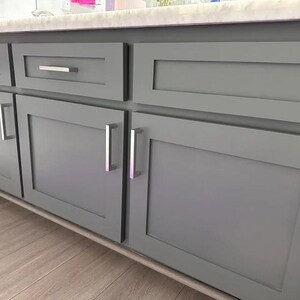 May include: A set of gray shaker-style cabinets featuring flat recessed center panels and rectangular silver metal handles. The cabinetry is topped with a light-colored stone countertop and includes a single drawer and two doors.