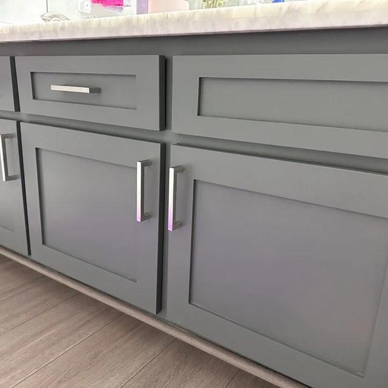 May include: A set of gray shaker-style cabinets featuring flat recessed center panels and rectangular silver metal handles. The cabinetry is topped with a light-colored stone countertop and includes a single drawer and two doors.