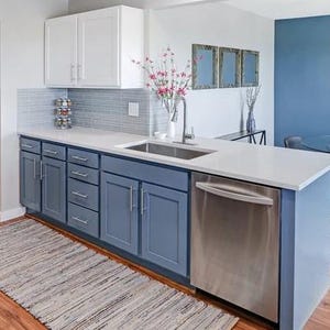 May include: A kitchen featuring white upper cabinets, blue lower cabinets, and a stainless steel dishwasher. The countertop is white with an integrated sink and faucet. A gray tiled backsplash sits above the counter, and a woven rug lies on the floor.