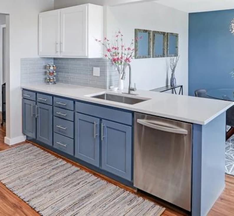 May include: A kitchen featuring white upper cabinets, blue lower cabinets, and a stainless steel dishwasher. The countertop is white with an integrated sink and faucet. A gray tiled backsplash sits above the counter, and a woven rug lies on the floor.