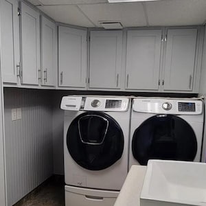 May include: A laundry room featuring light gray shaker-style cabinets with silver handles installed above two white Samsung front-load laundry machines with black circular doors. A white sink basin is visible in the foreground.