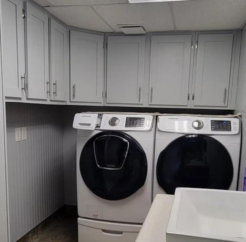May include: A laundry room featuring light gray shaker-style cabinets with silver handles installed above two white Samsung front-load laundry machines with black circular doors. A white sink basin is visible in the foreground.