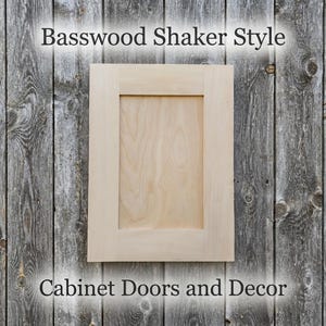 May include: A light-colored, unfinished basswood cabinet door with a flat center panel and a rectangular frame. The text Basswood Shaker Style and Cabinet Doors and Decor is visible above and below the wooden cabinet door.