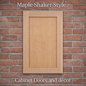 May include: A maple shaker style cabinet door against a red brick wall. The door is rectangular with a recessed panel and a light wood grain finish. The text "Cabinet Doors and décor" is at the bottom.