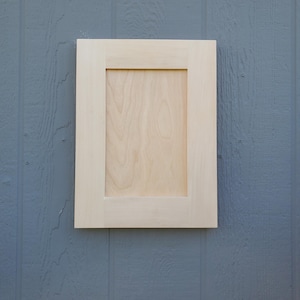 May include: A rectangular cabinet door made of light-colored, unfinished wood. The door features a flat, recessed center panel surrounded by a simple, wide frame with clean, straight edges and a smooth, natural wood grain texture.