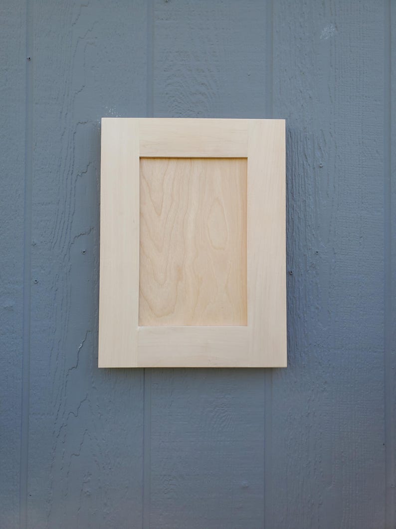 May include: A rectangular cabinet door made of light-colored, unfinished wood. The door features a flat, recessed center panel surrounded by a simple, wide frame with clean, straight edges and a smooth, natural wood grain texture.