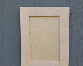 May include: A rectangular cabinet door made of light-colored, unfinished wood. The door features a flat, recessed center panel surrounded by a simple, four-piece frame with visible natural wood grain patterns throughout the surface.
