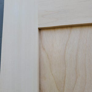 May include: A light-colored wooden cabinet door featuring a flat center panel and a wide frame. The wood grain is visible on the surface, showing natural light beige and cream tones with a smooth, unfinished texture throughout the entire piece.