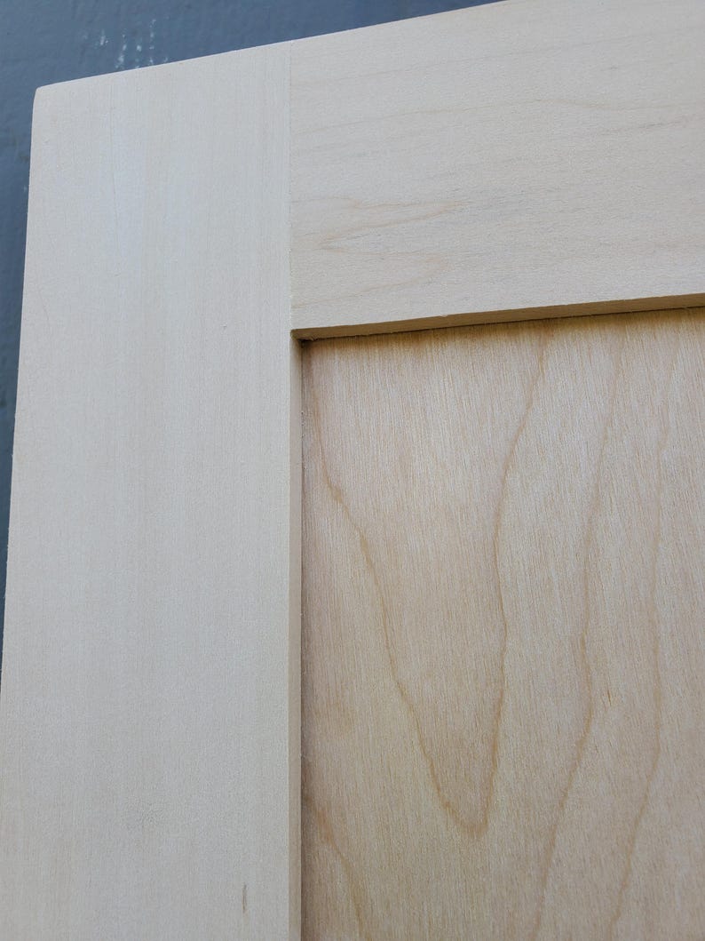 May include: A light-colored wooden cabinet door featuring a flat center panel and a wide frame. The wood grain is visible on the surface, showing natural light beige and cream tones with a smooth, unfinished texture throughout the entire piece.