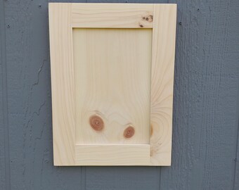 May include: A rectangular light-colored wooden cabinet door featuring a flat center panel and a surrounding frame. The natural wood grain is visible throughout, with two distinct circular knots located on the lower portion of the panel.
