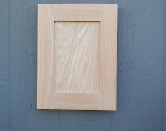 May include: A rectangular cabinet door made of light-colored, unfinished wood with a visible grain pattern. The door features a classic shaker-style design with a flat center panel surrounded by a simple, clean-lined frame border.