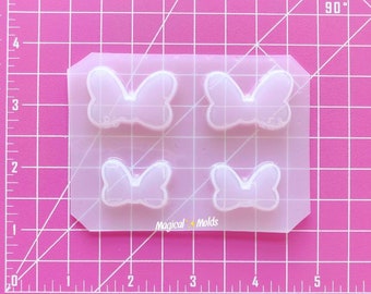 Puede incluir: Un molde de silicona transparente con cuatro cavidades en forma de lazos. El molde está sobre un fondo de rejilla rosa. El texto "Magical Molds" está impreso en el molde.