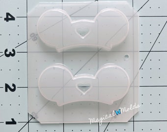 Puede incluir: Un molde de silicona transparente con dos cavidades en forma de orejas de Mickey Mouse con un corazón en el centro de cada oreja. El molde está sobre un fondo de cuadrícula blanca con líneas negras.