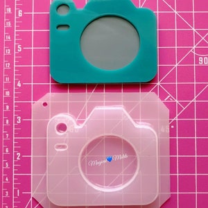May include: A clear silicone mold shaped like a camera. The mold is on a pink gridded surface. The text "Magical Molds" is visible in the mold.
