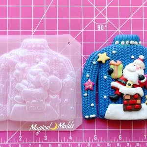 May include: A blue silicone mold for making a Christmas sweater ornament. The mold is shaped like a sweater with a Santa Claus design, a chimney, and stars. The mold is on a pink grid background.