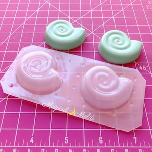 Op de afbeelding: Een roze siliconen mal met twee schelpenafdrukken en twee afgewerkte zeegroene schelpgietstukken. De mal heeft de tekst "Magical Molds". De achtergrond is een roze raster.