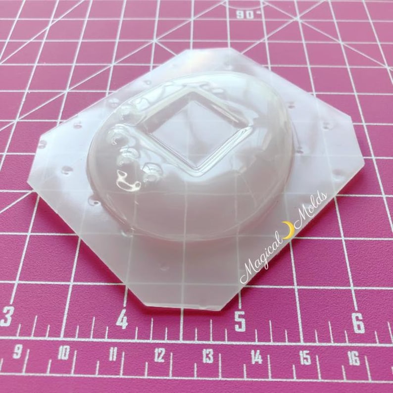 May include: Clear plastic mold for making resin crafts. The mold is shaped like a donut with a square cutout in the center. The mold is on a pink grid background.