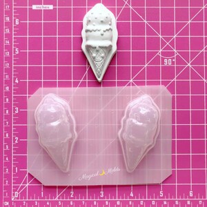 Puede incluir: Un molde de plástico transparente para hacer artesanías de resina con forma de cono de helado. El molde tiene dos cavidades y se muestra en una estera de corte rosa con una regla en pulgadas. El molde mide 6 pulgadas de ancho y 4 pulgadas de alto.