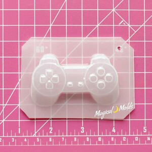 May include: A clear plastic mold of a video game controller. The mold is approximately 3 inches wide and 2 inches tall. The mold has a small hole at the top for hanging.