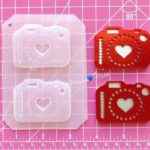 May include: Two clear silicone molds for making resin crafts. Each mold is shaped like a camera with a heart in the center. The molds are on a pink grid background.