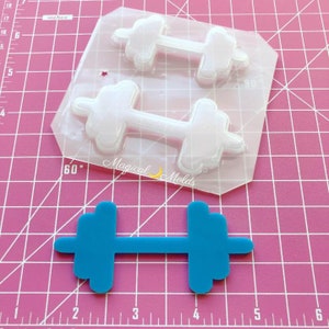 Op de afbeelding: Een transparante plastic mal voor het maken van twee halters. De mal ligt op een roze geruit oppervlak. Een blauwe halter ligt op de voorgrond.
