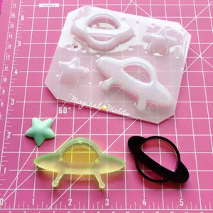 May include: A clear silicone mold with five different shapes: a spaceship, a planet with rings, a star, and two flying saucers. The mold is on a pink grid background. The text "Magical Molds" is visible on the mold.