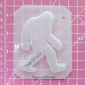 May include: Clear plastic mold of a Bigfoot, or Sasquatch, walking. The mold is 3.5 inches by 2.5 inches. The mold is on a pink grid background.