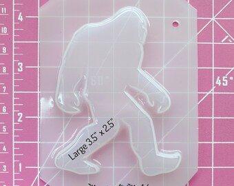 Könnte beinhalten: Transparente Plastikform eines Bigfoot oder Sasquatch beim Gehen. Die Form ist 3,5 Zoll mal 2,5 Zoll groß. Die Form befindet sich auf einem rosa Gitterhintergrund.