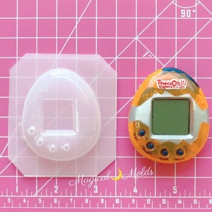 May include: A clear plastic mold shaped like a Tamagotchi, a handheld digital pet, with a square screen area and four small circles around the bottom. Next to the mold is a yellow and orange Tamagotchi Connection digital pet with a screen that reads "Tamagotchi Connection".
