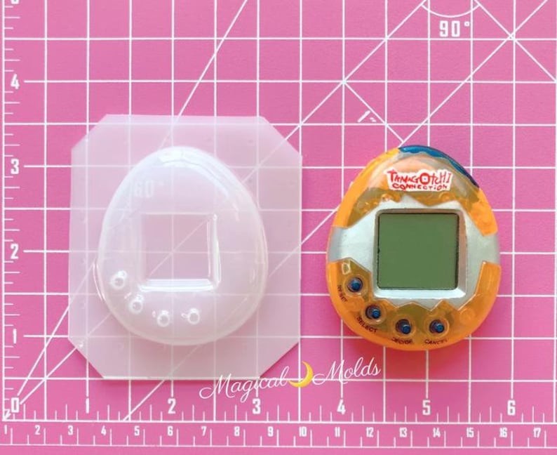 May include: A clear plastic mold shaped like a Tamagotchi, a handheld digital pet, with a square screen area and four small circles around the bottom. Next to the mold is a yellow and orange Tamagotchi Connection digital pet with a screen that reads "Tamagotchi Connection".