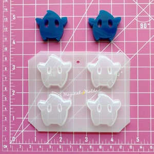 May include: A clear silicone mould with four star-shaped cavities. Each cavity has a design resembling a video game character with two eyes and a mouth. Two blue resin castings of the same design are shown next to the mould.