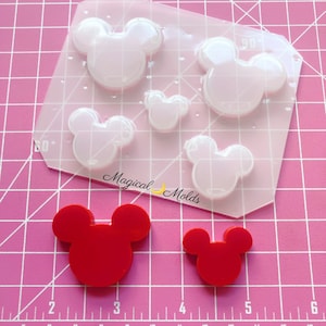 May include: A clear plastic mold with five Mickey Mouse head shapes. Two of the shapes are large, two are medium, and one is small. The mold is on a pink gridded surface. There are two red Mickey Mouse head shapes on the surface, one large and one small.