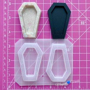 May include: A clear silicone mould with two coffin-shaped cavities. The mould is on a pink gridded surface. There are two finished resin pieces, one black and one with glitter, in the shape of a coffin.