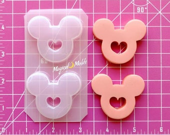 Puede incluir: Dos moldes de silicona transparentes para hacer manualidades de resina. Cada molde tiene la forma de una cabeza de Mickey Mouse con un corazón recortado en el centro. Los moldes están sobre un fondo de rejilla rosa.