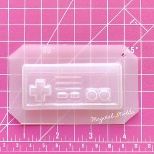 May include: A clear, rectangular silicone mold featuring a retro video game controller design. The mold includes a directional pad, buttons, and a logo that reads "Magical Molds". The mold is on a pink grid background.