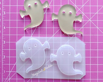 May include: Clear, ghost-shaped molds and finished products are displayed on a pink grid background. The molds are in a rectangular shape with the text "Magical Molds". The finished ghosts are approximately 2 inches tall.