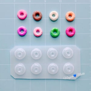 May include: A collection of colorful miniature donuts in pink, brown, white, and orange, alongside a clear silicone mold with donut-shaped cavities. The background is a light blue surface with grid lines.