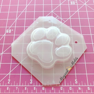 Könnte beinhalten: Transparente Plastikform in Form eines Pfotenabdrucks. Die Form befindet sich auf einer rosa karierten Oberfläche. Der Text "Magical Molds" befindet sich auf der Form.