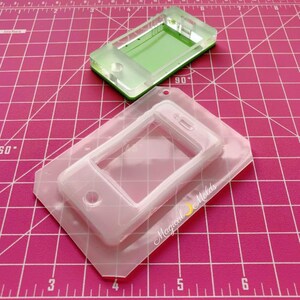 May include: Clear silicone mold for making phone-shaped resin crafts. The mold is shaped like a smartphone with a rectangular screen and a round button at the bottom. The mold is on a pink cutting mat.