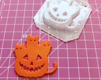 May include: Orange silicone mold of a pumpkin with three cats on top. The mold is on a pink cutting mat with a ruler.