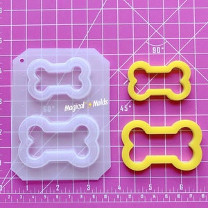 May include: A clear plastic mold with two bone-shaped cavities. The mold is on a pink cutting mat with white markings. There are two yellow bone-shaped silicone molds next to the plastic mold. The text "Magical Molds" is printed on the plastic mold.