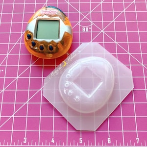 May include: A clear plastic mold for making a replica of a Tamagotchi toy. The mold is shaped like a teardrop and has a square-shaped hole in the center. The mold is sitting on a pink gridded surface.