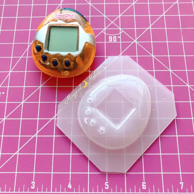 May include: A clear plastic mold for making a replica of a Tamagotchi toy. The mold is shaped like a teardrop and has a square-shaped hole in the center. The mold is sitting on a pink gridded surface.