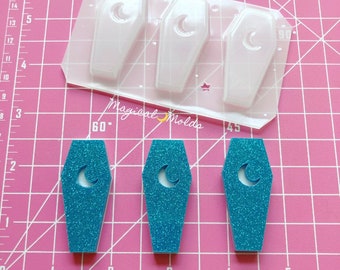 May include: Three clear plastic molds shaped like coffins with a crescent moon design. Three teal blue glitter filled coffins with crescent moon designs are shown on a pink cutting mat.