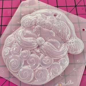 May include: Clear plastic mold for making a Santa Claus ornament. The mold features a detailed design of Santa's face with a curly beard and a hat. The mold is on a pink surface with a white grid pattern. The text "Magical Molds" is visible in the bottom right corner.