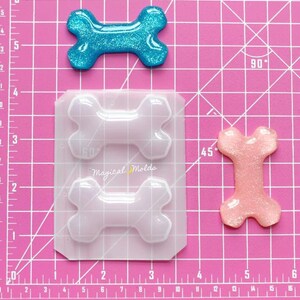 Puede incluir: Un molde de silicona transparente con dos cavidades en forma de hueso. El molde está sobre una superficie cuadriculada rosa. Hay dos piezas de resina terminadas en forma de hueso, una azul y una rosa, con purpurina incrustada en la resina.
