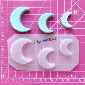 Peut inclure: Un moule en plastique transparent avec quatre formes de croissant de lune. Le moule est sur un fond de grille rose. Le moule est étiqueté "Magical Molds" et comporte des marques de degrés de 45 et 90 degrés.