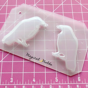 May include: Clear plastic mold for making two resin crow figurines. The mold is on a pink gridded surface. The text "Magical Molds" is visible on the mold.