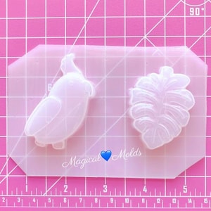 May include: A clear silicone mold with two designs: a bird and a tropical leaf. The mold is on a pink gridded surface.